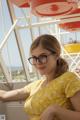 A woman in a yellow dress and glasses sitting on a ferris wheel.